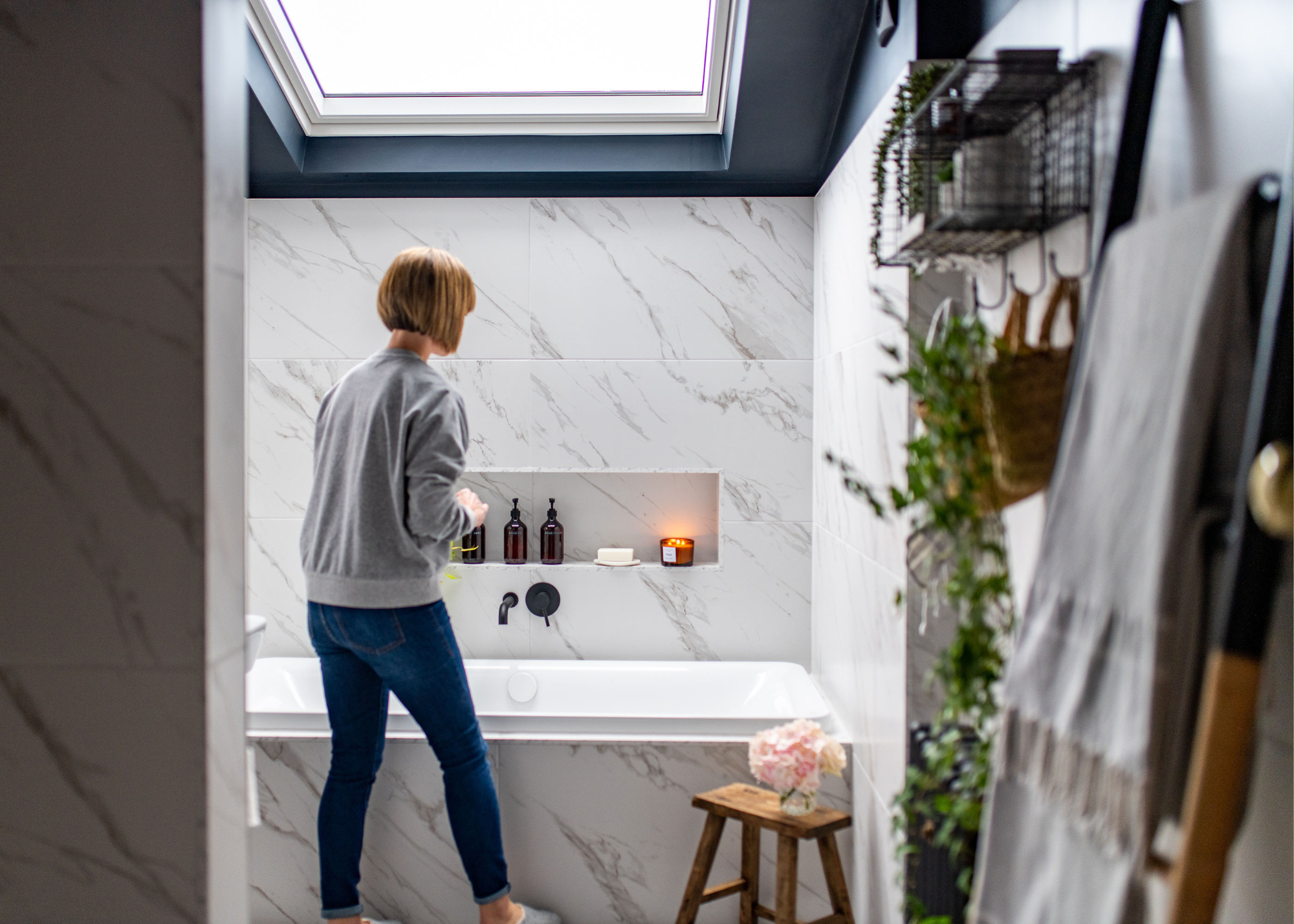 A stone bathroom with a woman with a short bob in a gray sweater and skinny jeans near a bathtub, under a roof window letting light int