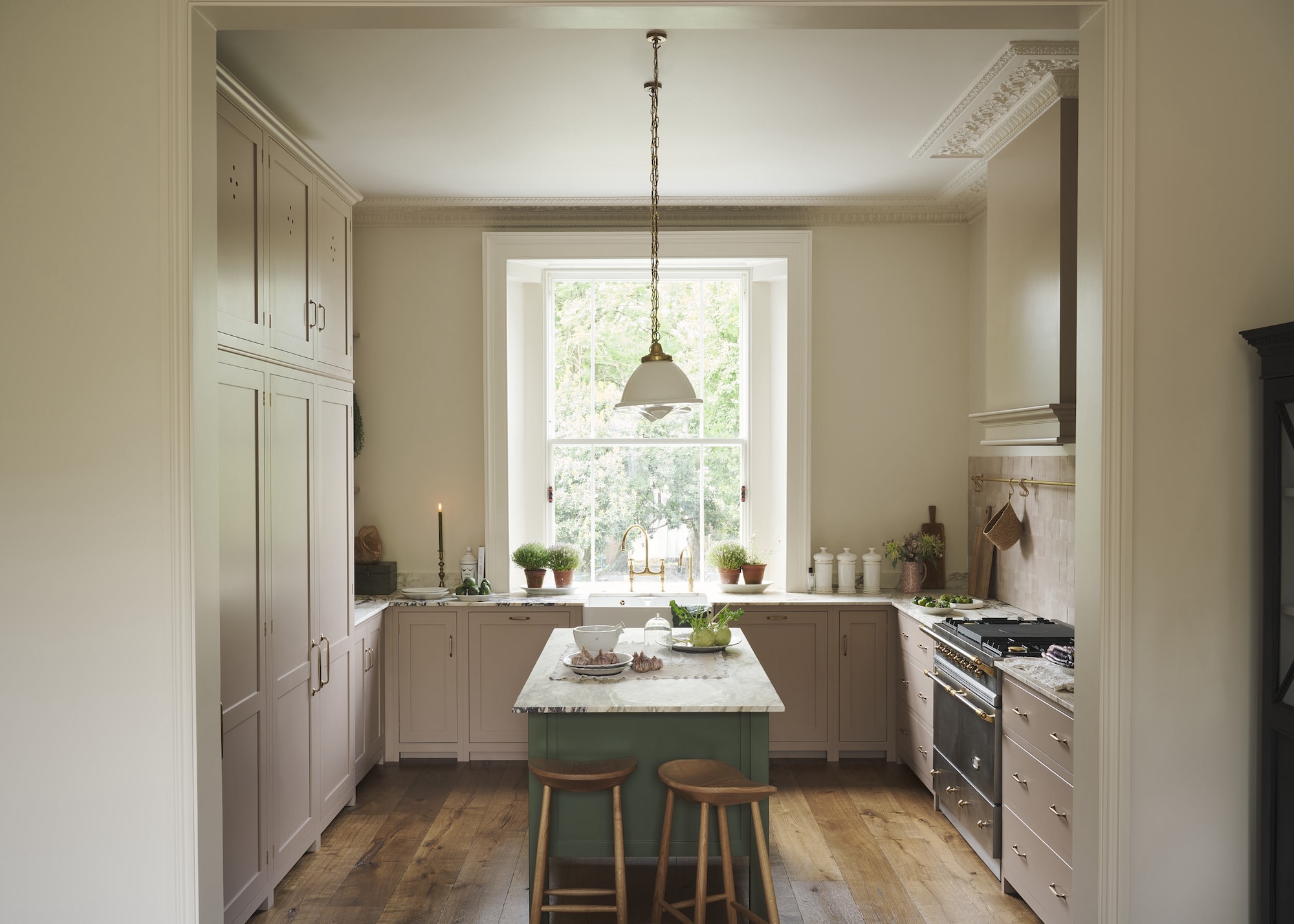 A kitchen with a green colored small island in the middle. Surrounding the island are baby pink cabinets. Above the island is a hanging pendant light.