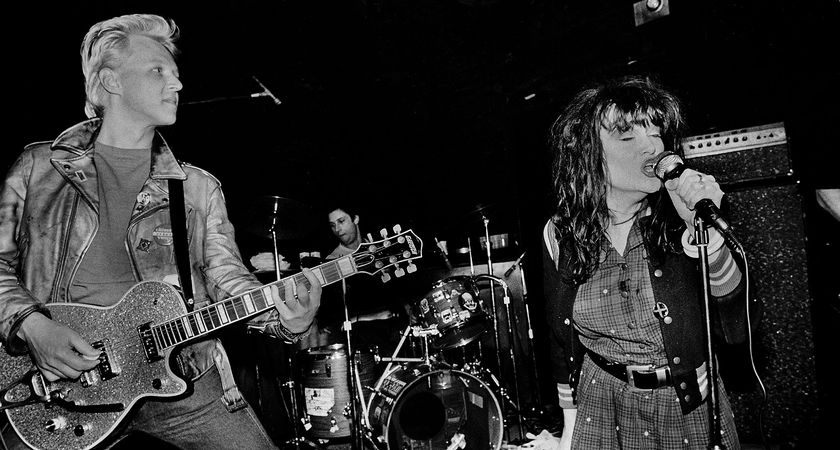 A black-and-white action shot of Billy Zoom of X playing rhythm on his Gretsch as L.A. punks X tear it up onstage. On the right of the picture is vocalist Exene Cervenka.