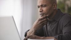 A man appears to be worried as he looks at his laptop at his dining room table.