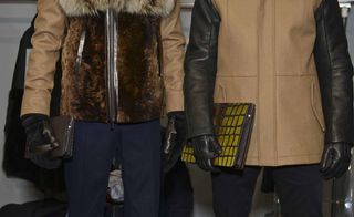 Two male models wearing looks from Fendi's collection. They are wearing blue trousers with one model wearing a brown leather and fur jacket and the other model wearing a brown and black leather coat. Both models are holding rectangular bags with handles