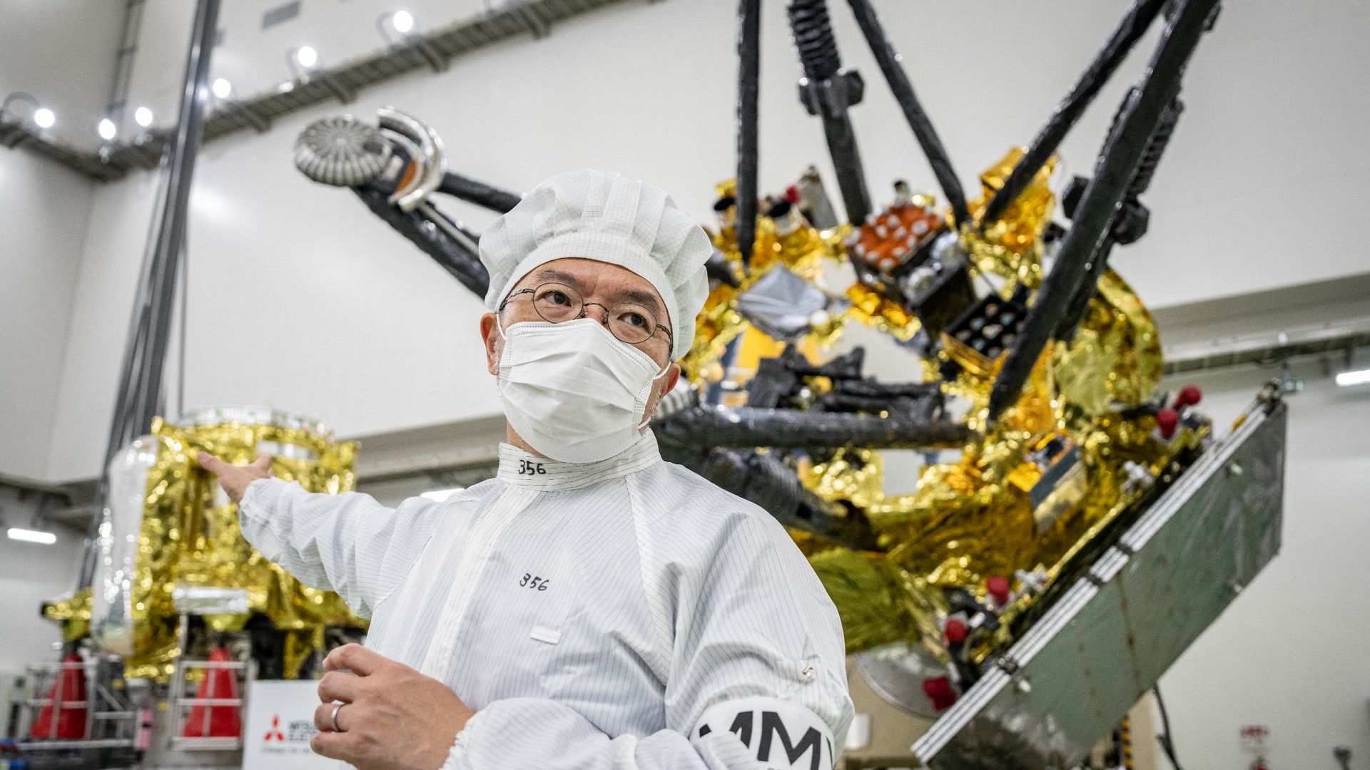 A photo of a man in white coveralls and a face mask stood in front two parts of the MMX probe in a clean room