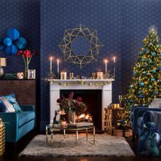 Dark blue livng room, Christmas tree with Christmas decorations, white fireplace, lit fire, gold coffee table, blue sofas with patterned cushions.