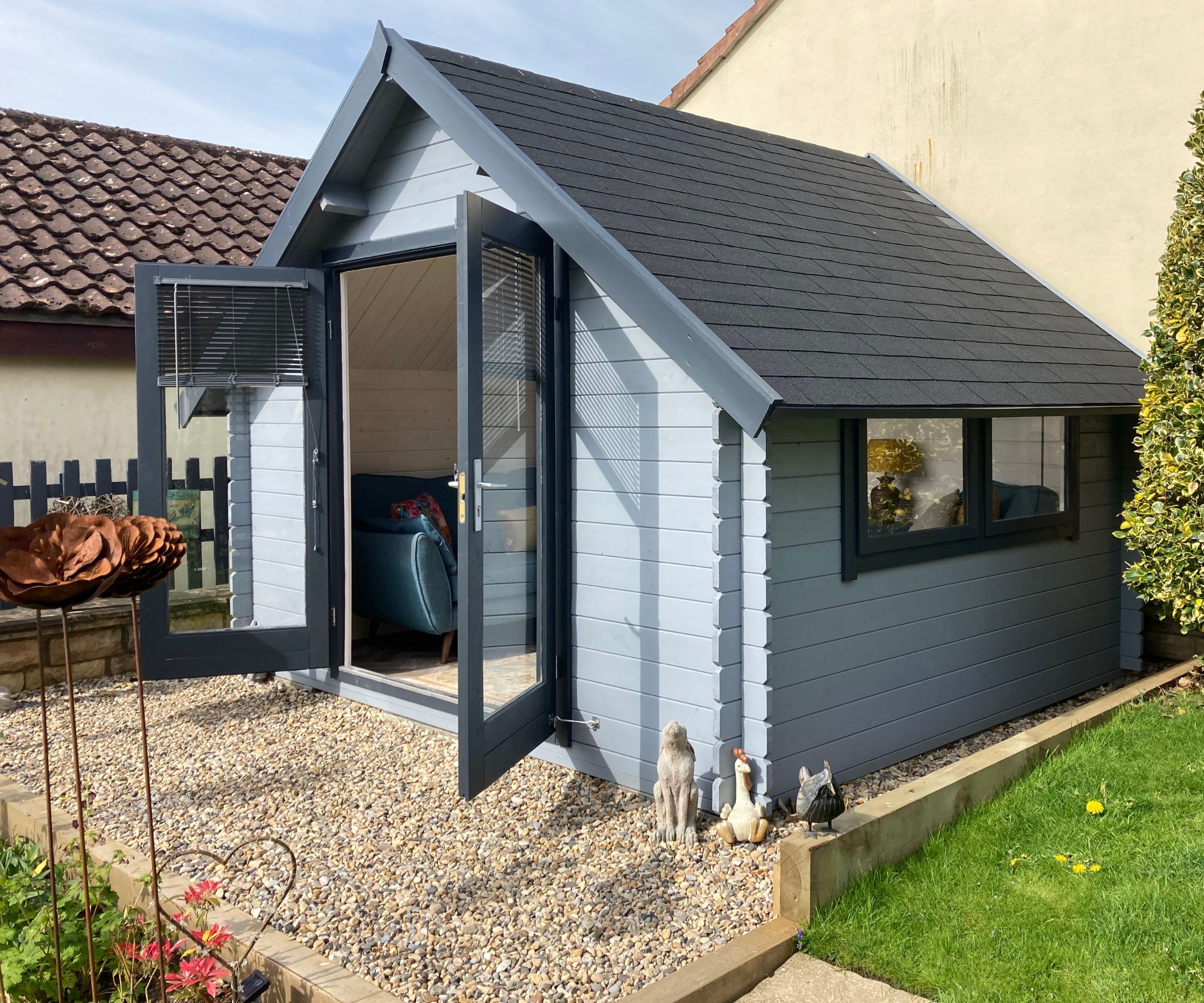 A grey painted apex-roofed garden shed with dark shingle roof, open glazed double doors revealing a teal sofa inside, on a gravel base with animal ornaments