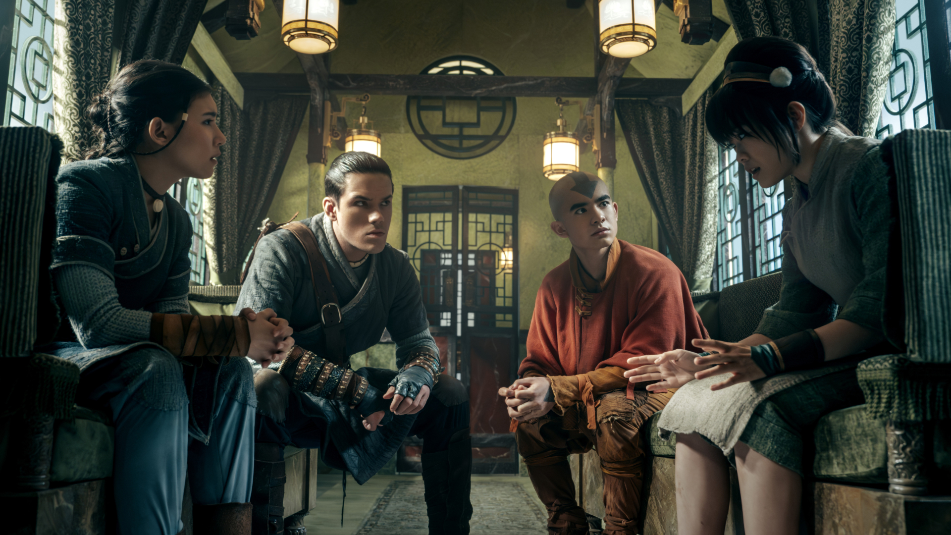 A group of kids (l-r Kiawentiio Tarbell as Katara, Ian Ousley as Sokka, Gordan Cormier as Aang, Miya Cech as Toph) sit in a room decorated with metal encircled squares, in 'Avatar: The Last Airbender' season 2.