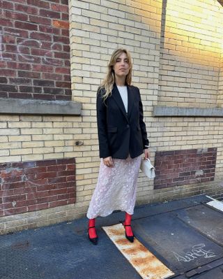 A woman stands against a brick wall wearing a black blazer with a white lace skirt, red tights, and black pointy toe heels.