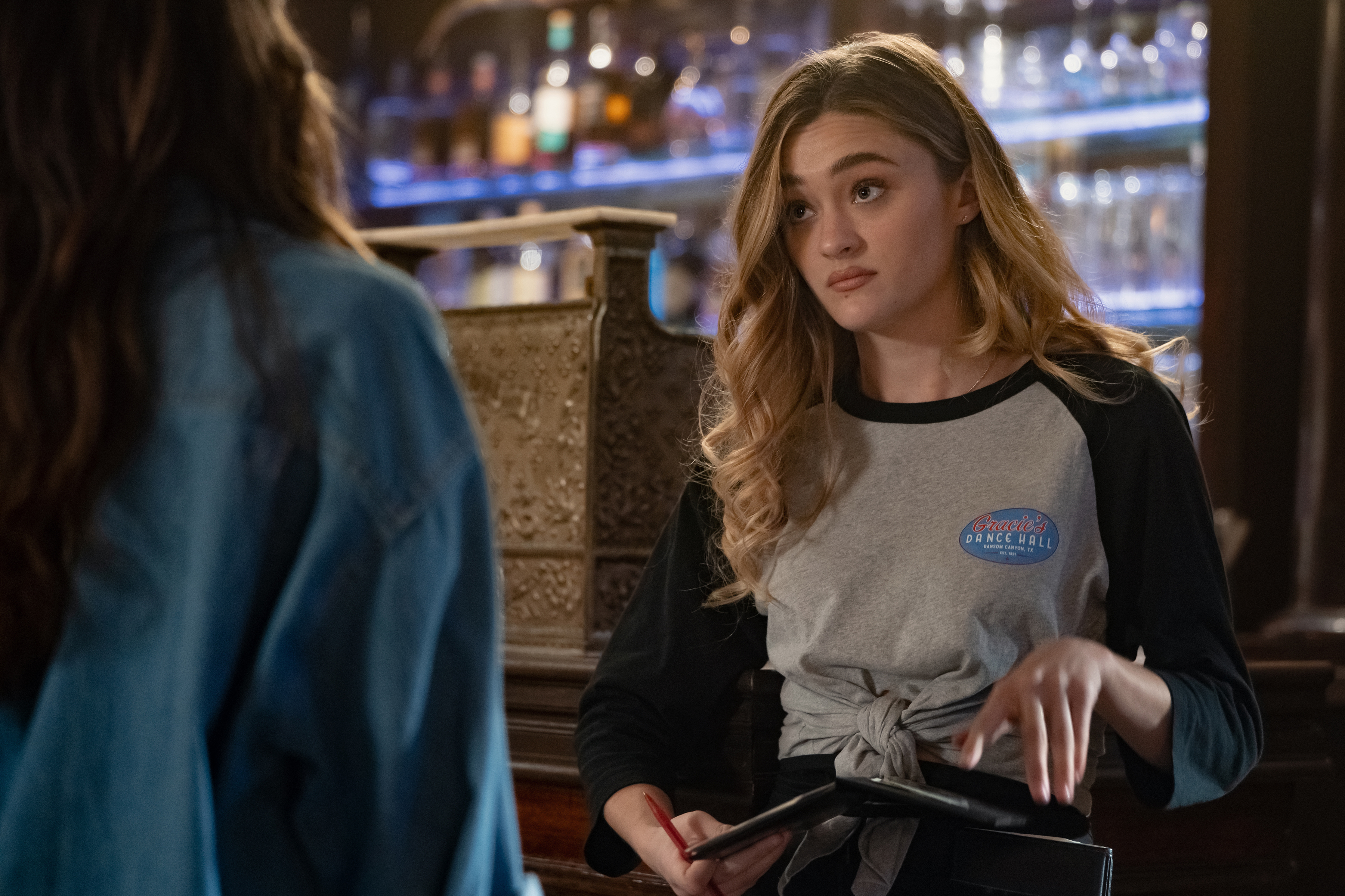 Lizzy Greene as Lauren holds a bill holder while standing in a restaurant, in Episode 203 of Netflix's 'Ransom Canyon' Season 2.