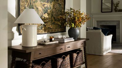 entryway table from the mcgee & co fall collection styled with a bobbin vintage style console table with a tapestry hung behind and styled with baskets, a lamp and vase with foliage