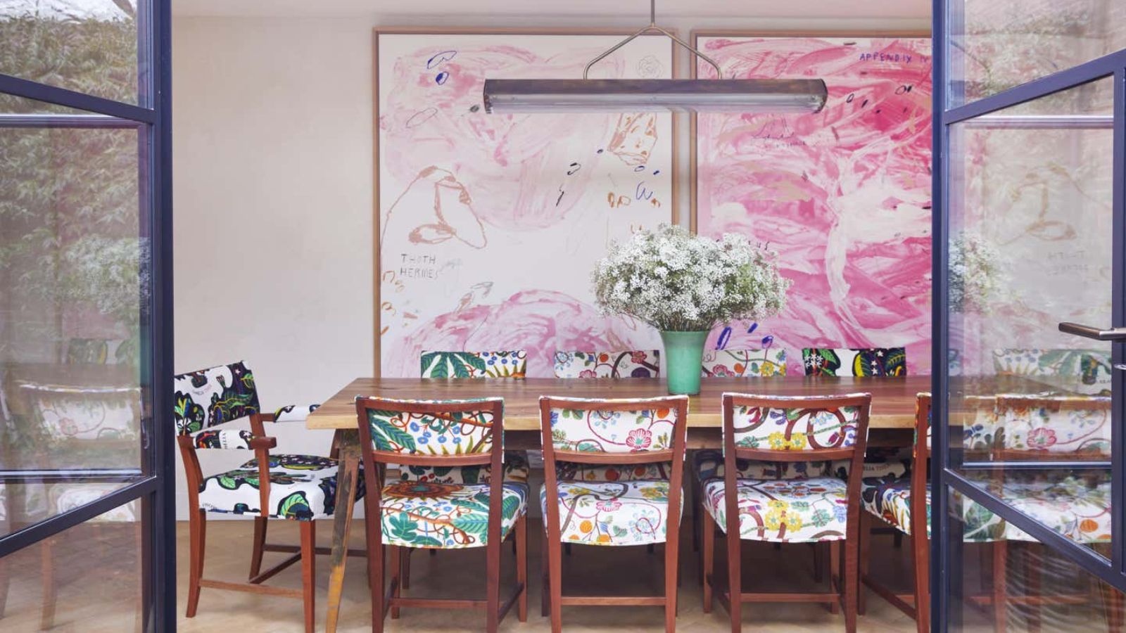 A dining room with large pink swirl artwork on the walls. A long wooden rectangular dining table with colorful chairs around it. A large green vase of white flowers as a centrepiece. 