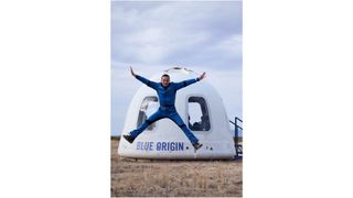 a man in a blue flight suits does a star jump in front of his white space capsule after landing on a suborbital flight.