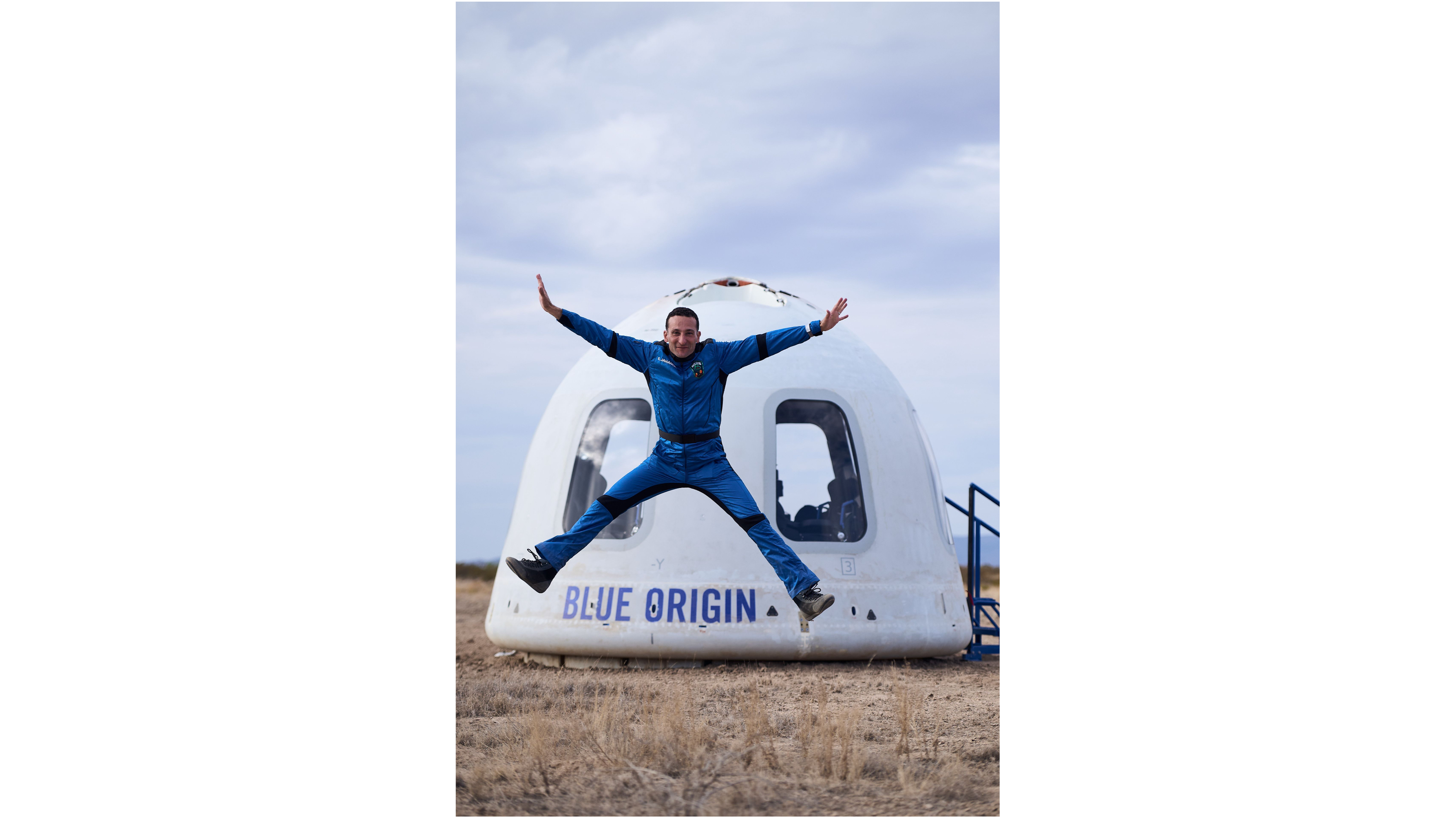 a man in a blue flight suits does a star jump in front of his white space capsule after landing on a suborbital flight.