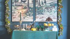 Console table with green tablecloth and drinks and treats on tiered stands with festive garland and painting on the wall behind