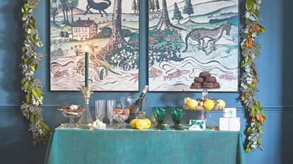 Console table with green tablecloth and drinks and treats on tiered stands with festive garland and painting on the wall behind