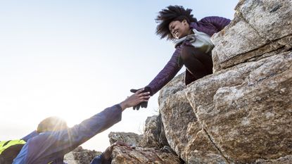 A hiker reaches down to help another hiker up a cliff.