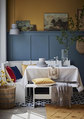 dining area with white bench seating, wooden floor, rug, fall color palette, panelled blue wall behind, baskets, velvet cushions in yellow, blue and red