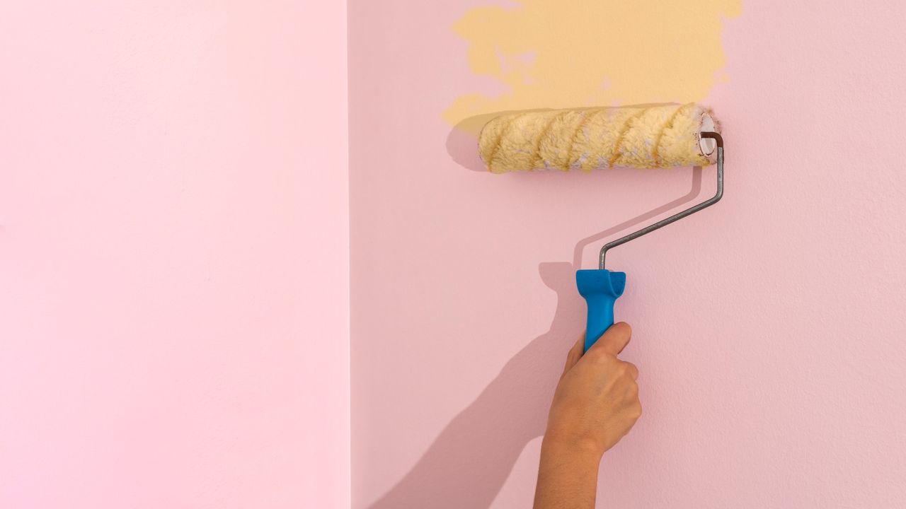 Cropped woman&#039;s hand painting a wall with a roller applying yellow colored paint to pink wall