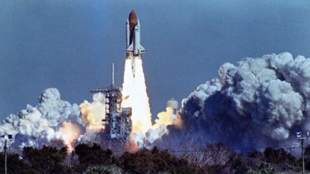 a white aircraft attached to a large orange rocket lifts off above a plume of fire and smoke