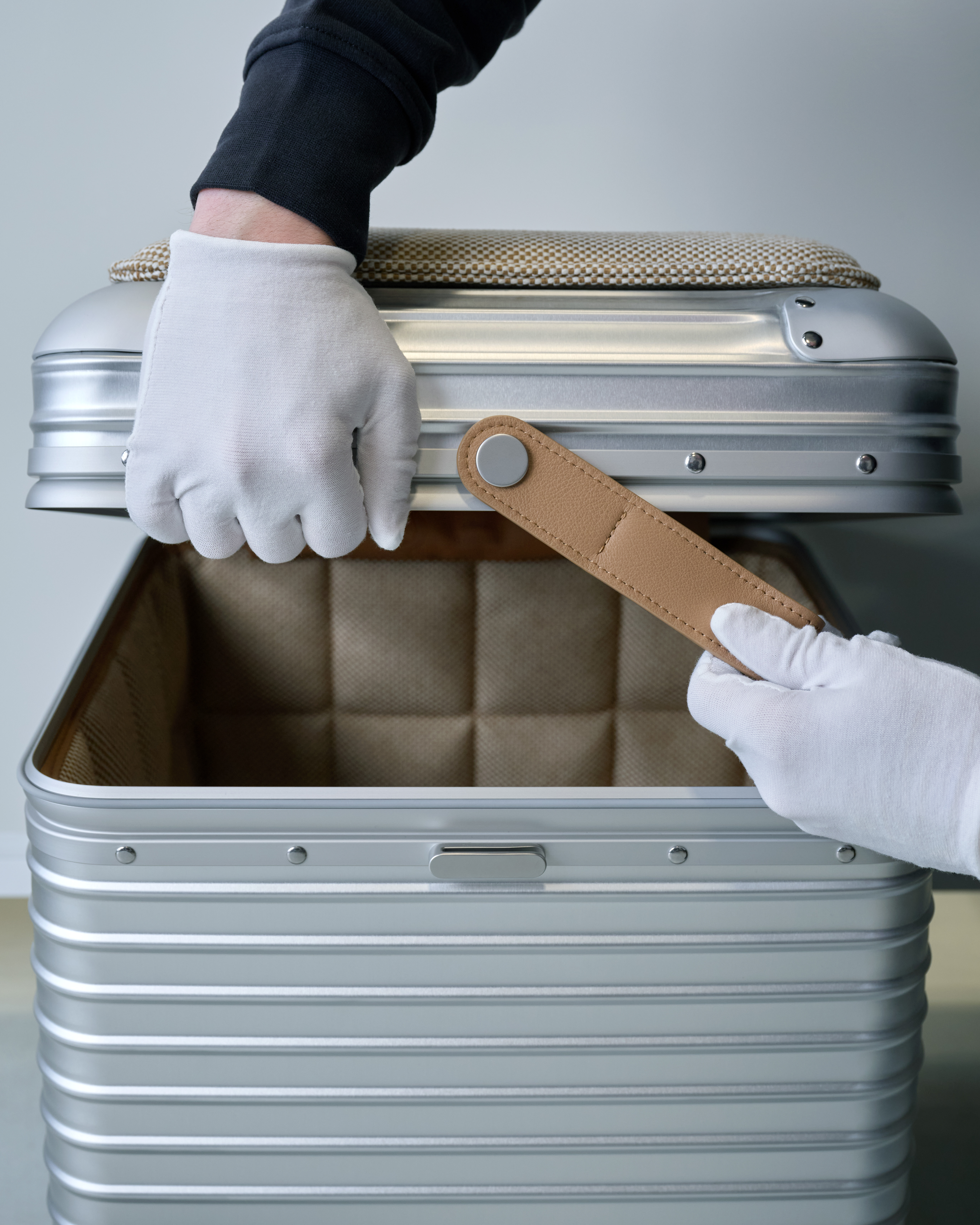 Close-up shot of white-gloved hands opening the RIMOWA x Vitra Aluminum Stool, revealing its beige leather tag and padded lining
