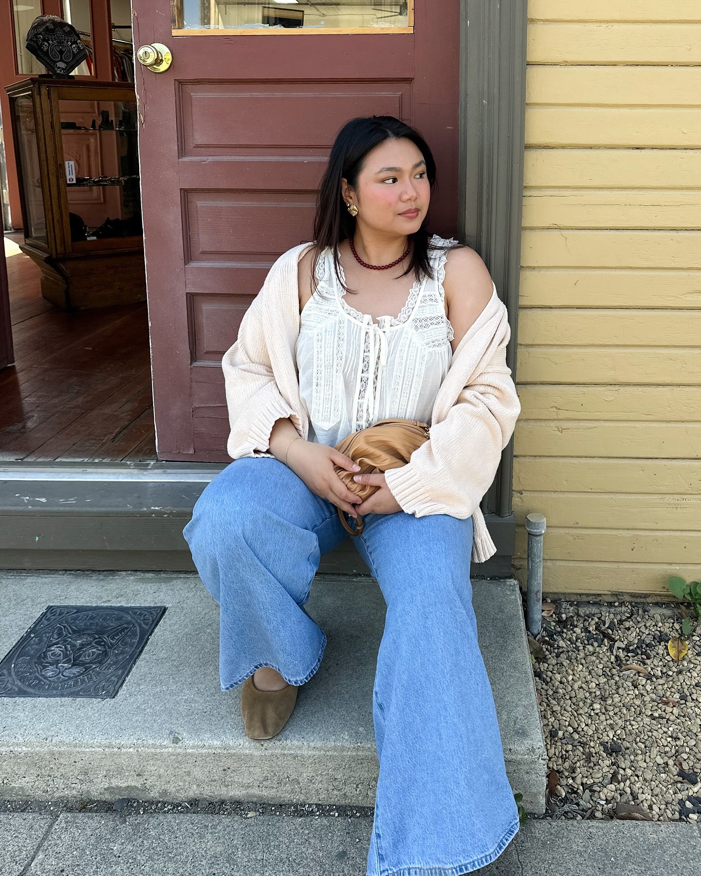 Fashion person Marina is wearing the lace camisole top trend, she pairs with a pink cardigan blue denim jeans, brown suede shoes and a light brown clutch with isn't to dissimilar in colour to her bag. She is sat in a doorway in front of a red - brown door in an image taken from her Instagram recently.