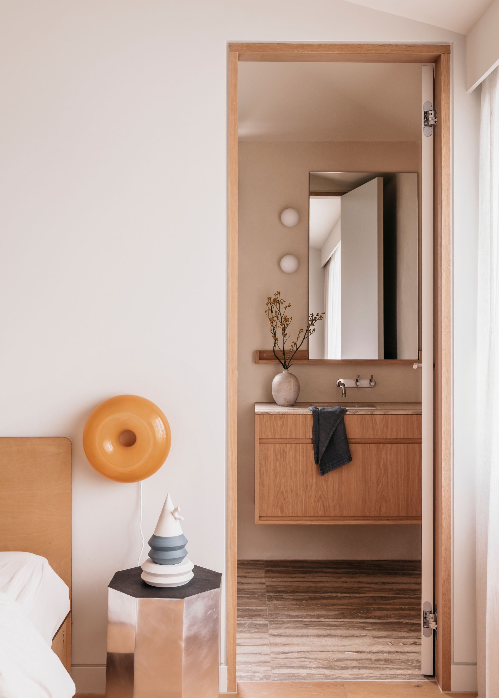 A white bedroom with an orange IKEA VARMBLIXT donut lamp on the wall, a steel side table, and a peek into a minimalist bathroom