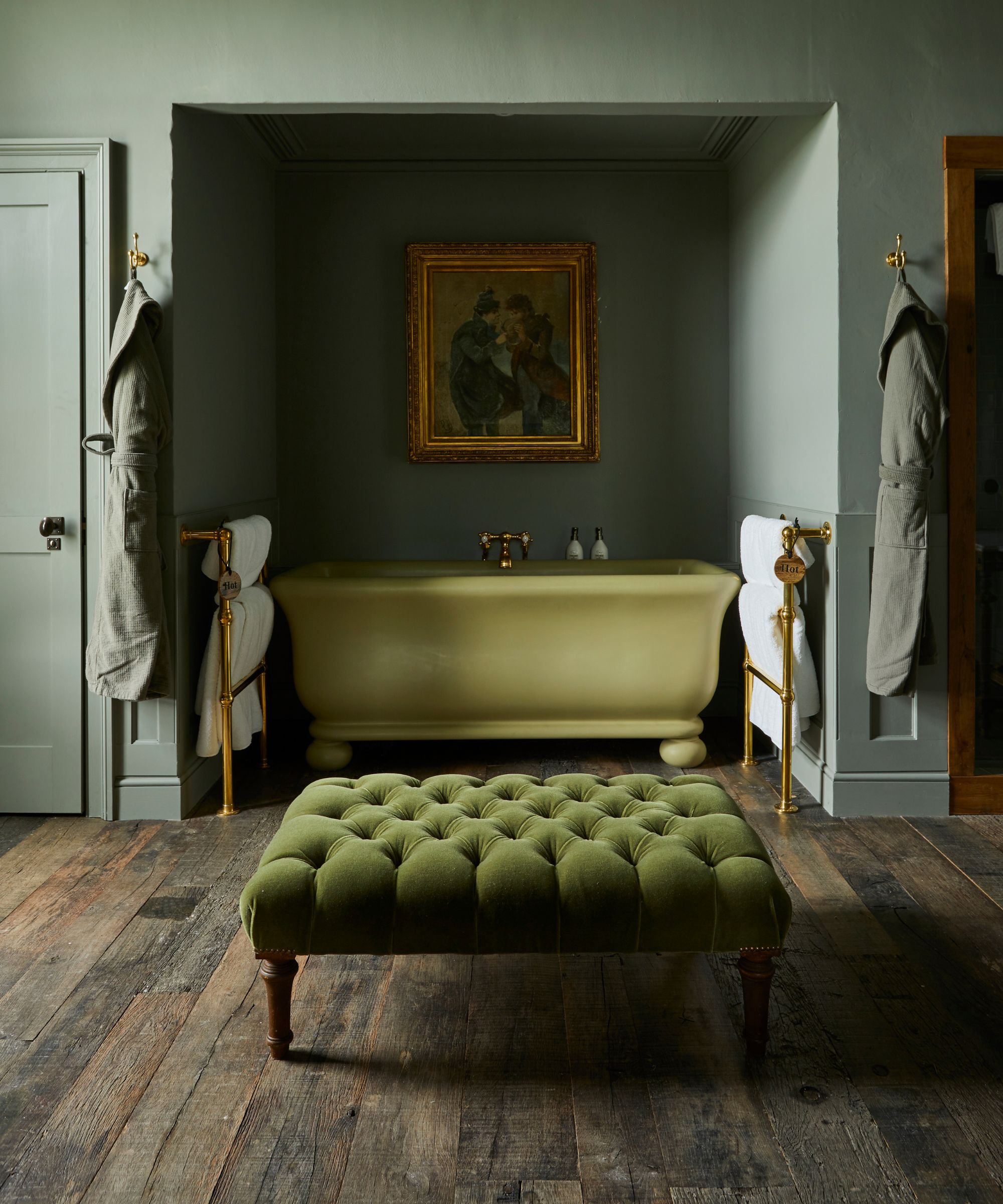 An olive green bathtub sitting in an alcove in the wall, with a large painting above in earthy tones with a gold frame. In the foreground is a dark green ottoman. The walls are a deep green colour and the floors are exposed floorboards.