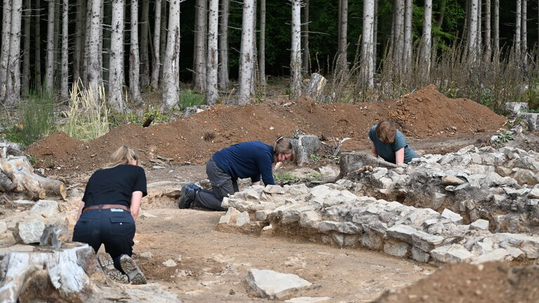 three light-skinned archaeologists kneel on the ground and dig near low stone walls