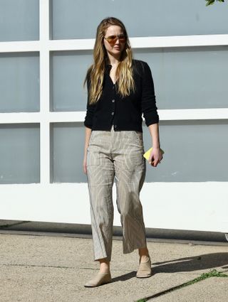 Jennifer Lawrence mengenakan kardigan hitam, celana panjang bergaris, dan sepatu flat The Row berbahan mesh.
