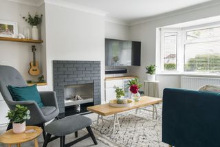 White living room with grey carpet, boho rug, grey armchair and footstool, wood coffee table, blue velvet sofa and grey brick fireplace surround