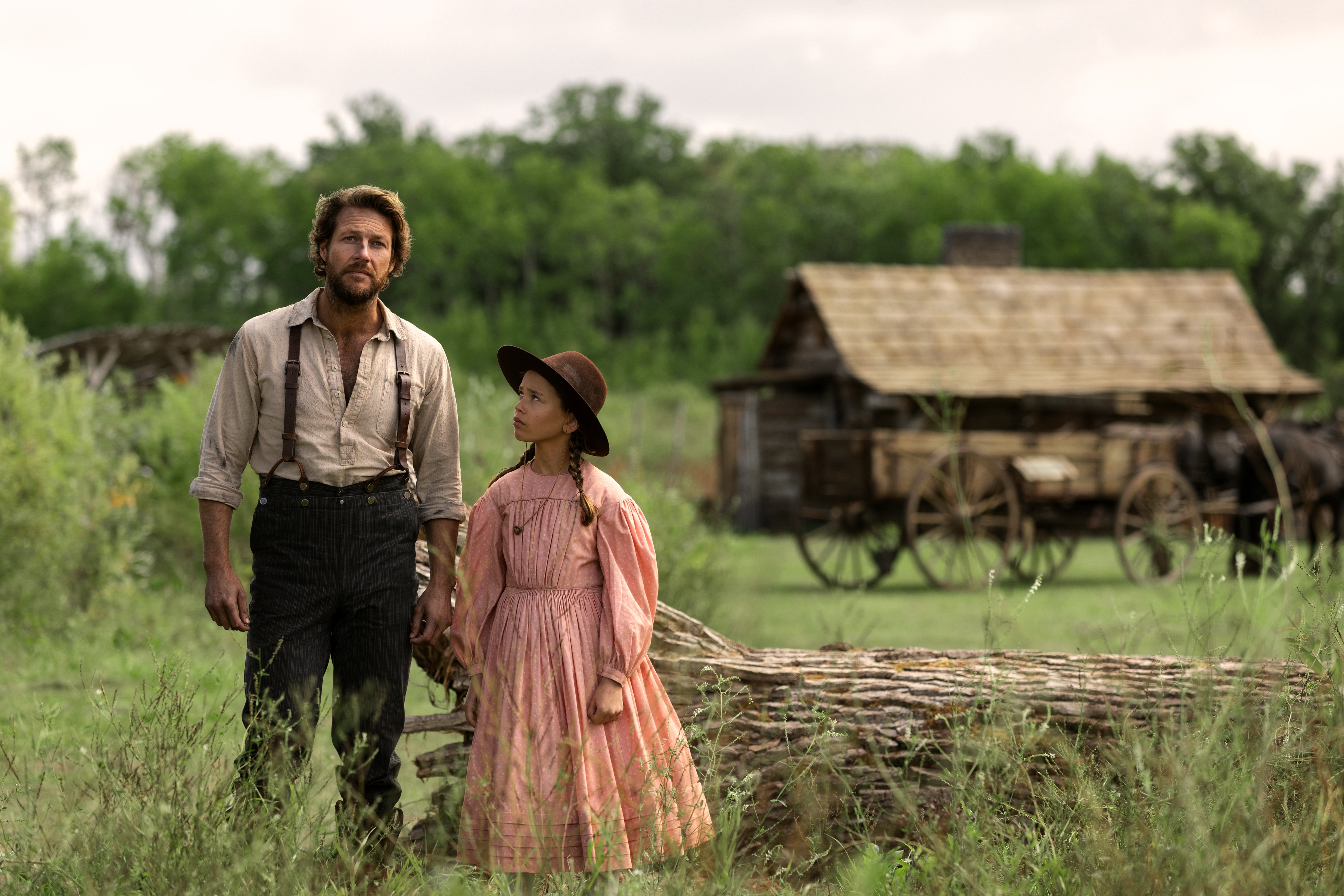Luke Bracey as Charles Ingalls and his daughter Alice Halsey as Laura Ingalls hold hands while standing in front of a frontier-era wagon and cart, in Netflix's 'Little House on the Prairie.'