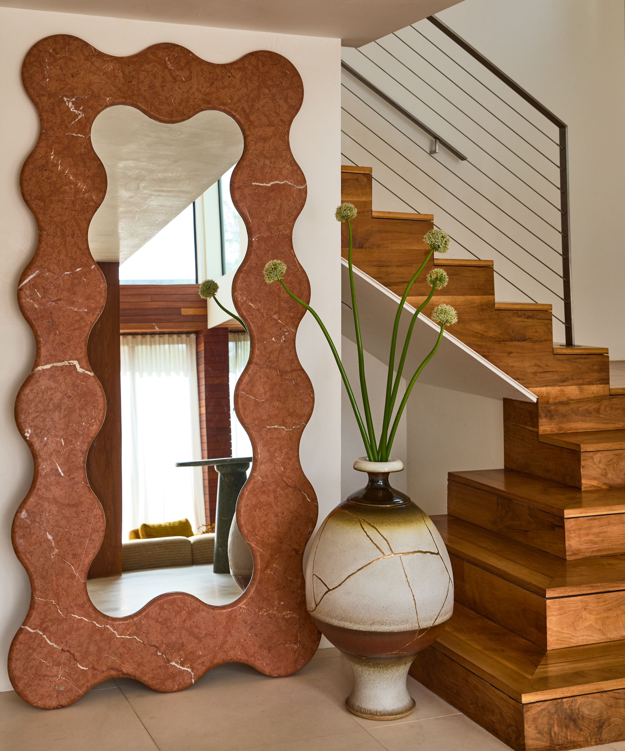 entryway stairs with tiled floor, large wavy red travertine mirror, and a large floor vase with sculptural stems in it and timber stairs