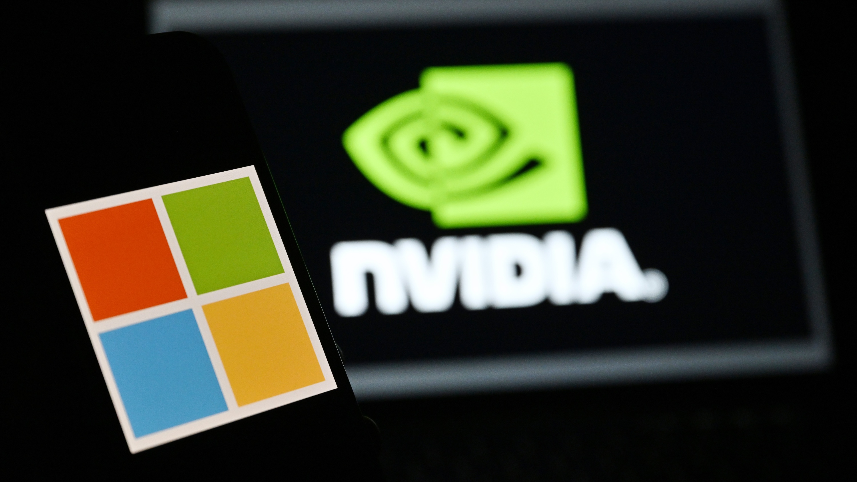 The &#039;Microsoft&#039; logo is displayed on a mobile phone screen in front of a computer screen displaying Nvidia logo.