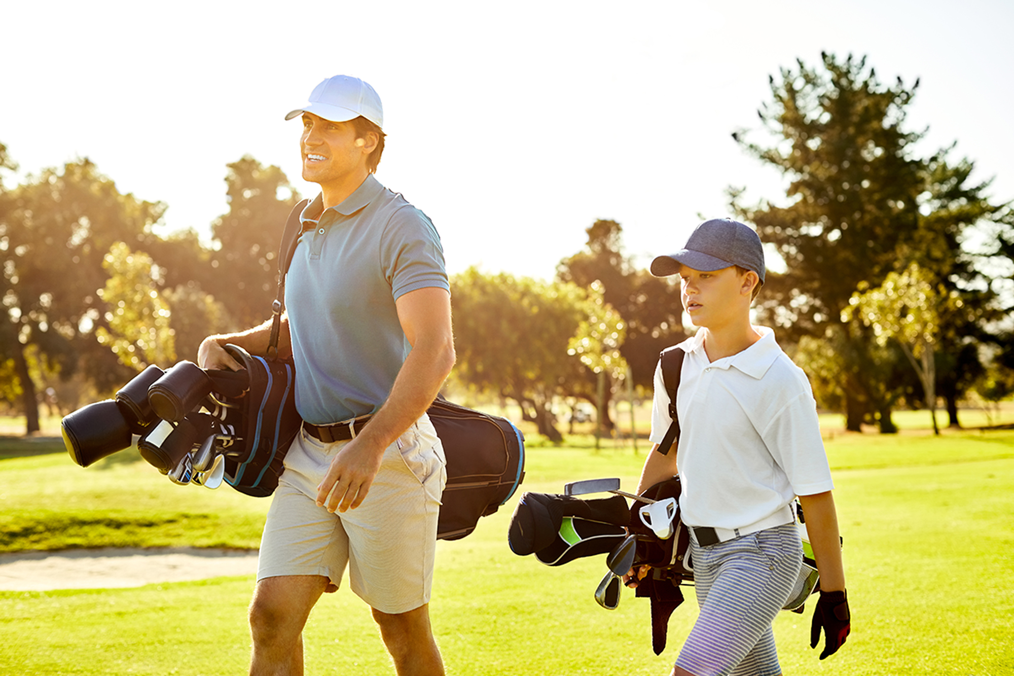 a father and son on the golf course, both dressed in traditional golf attire with a set of golf clubs looped over each of their shoulders