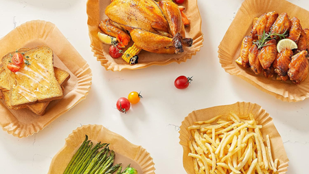A set of five square paper air fryer liners on a white countertop, viewed from above. A different food in each, including a toasted sandwich, a roast chicken, baked fish, asparagus, and fries. Three small tomatoes lay on the worktop between them. 