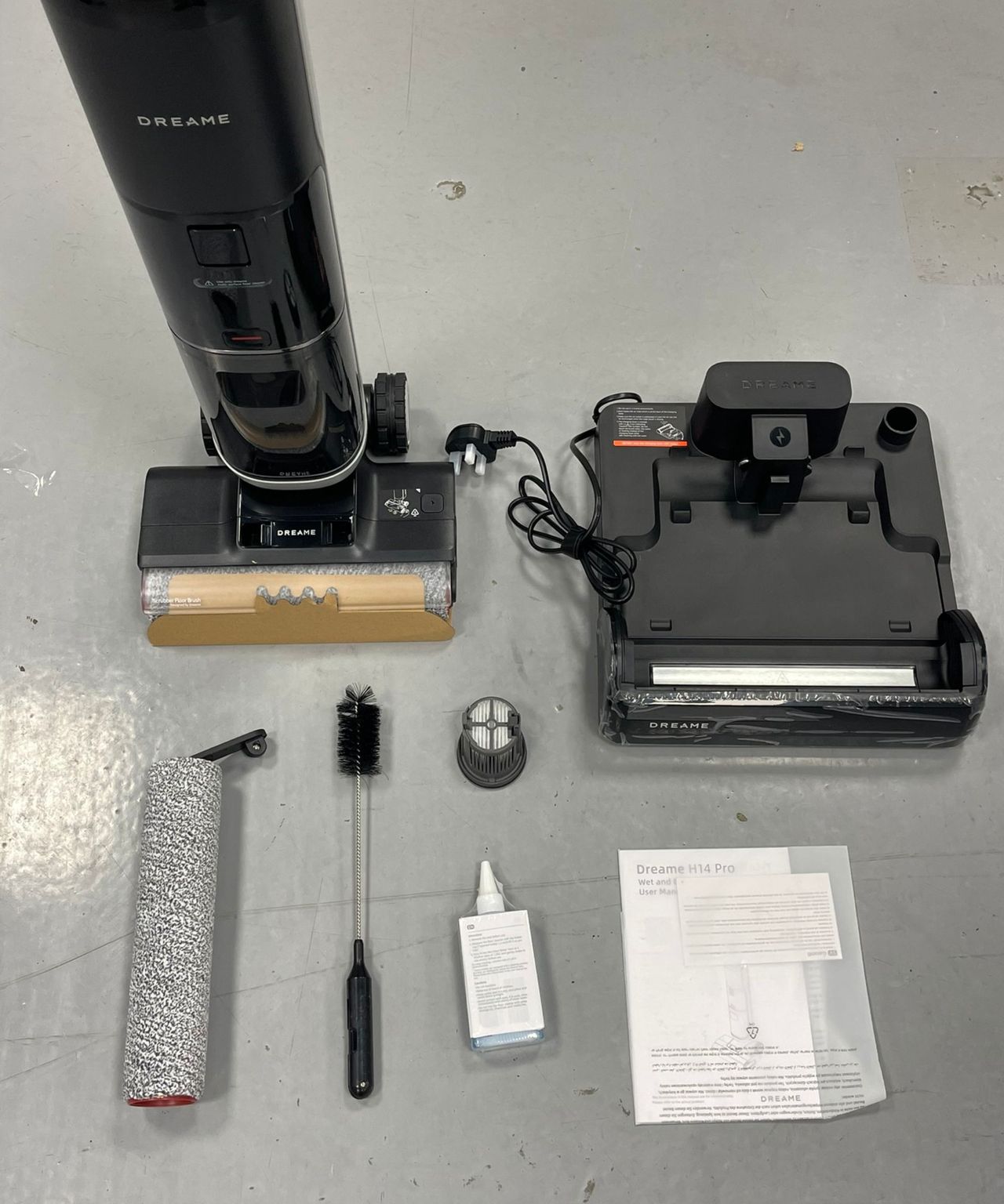 The Dreame H14 Pro vacuum and charging base, roller, cleaning brush, cleaning solution, filter and instruction manual laid out on gray floor in Future test center