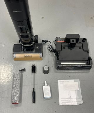 The Dreame H14 Pro vacuum and charging base, roller, cleaning brush, cleaning solution, filter and instruction manual laid out on gray floor in Future's test center