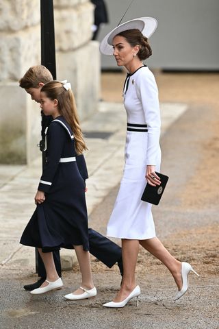 Kate Middleton and Prince Charlotte dressed in white and navy dresses and white shoes walking through wet gravel with Prince George in the background