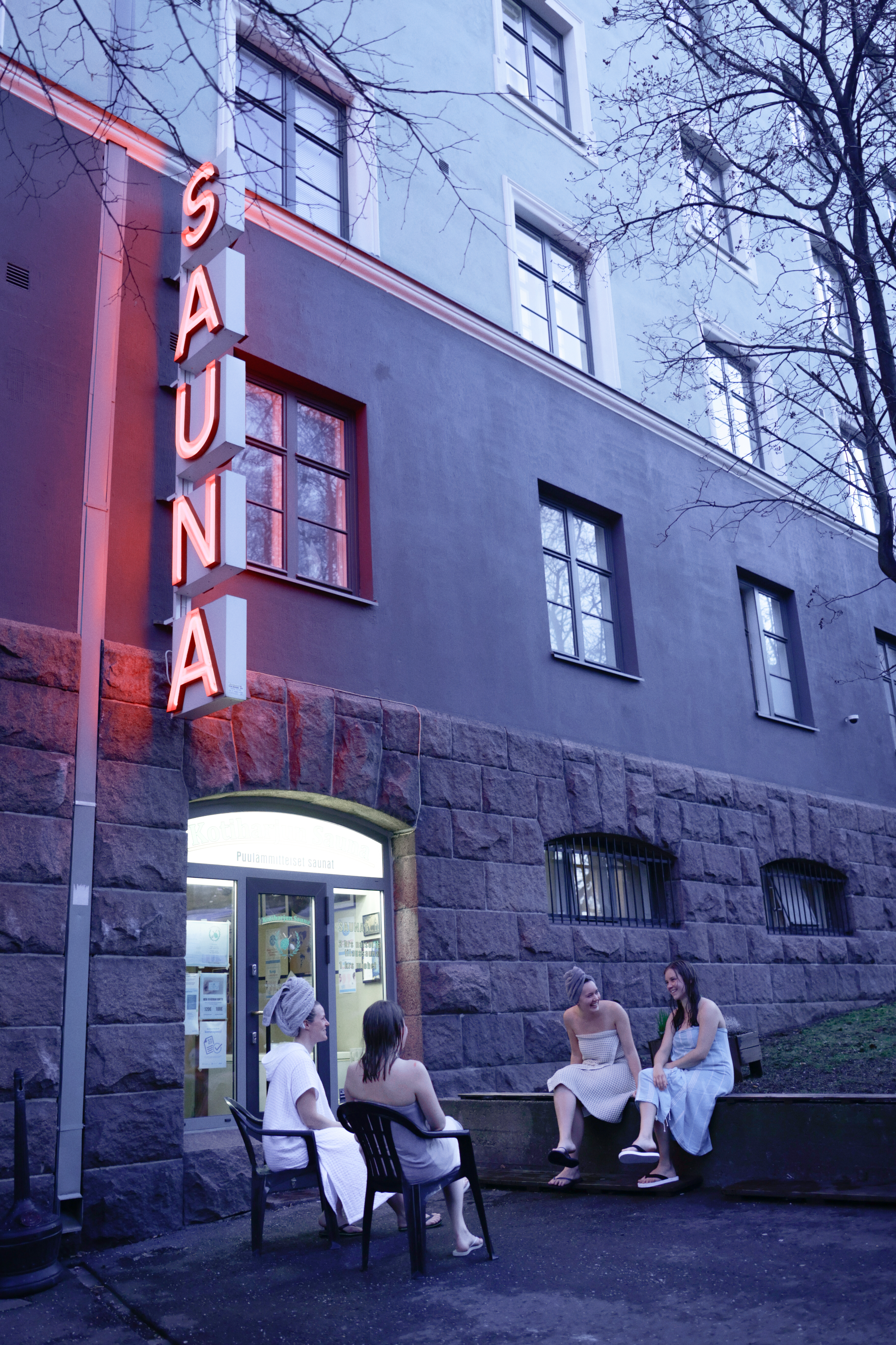Four young women wrapped up in long shower towels with their hair still wet sit outside a sauna whose sign features neon-lit, red lighting on the cusp of the evening.