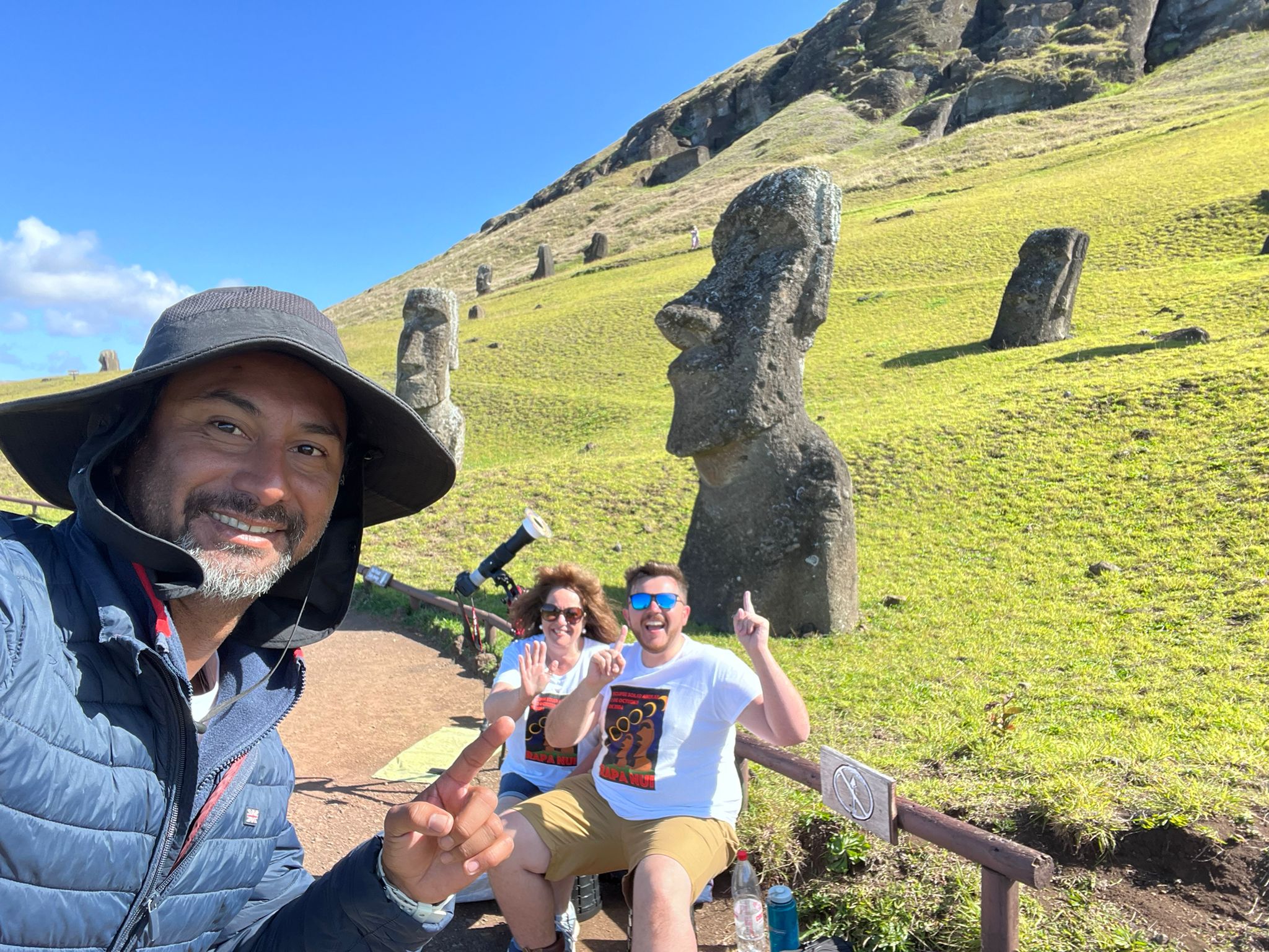 three people smiling with moai statues behind and lush green grass.