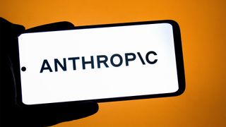 Anthropic logo and branding pictured on a smartphone held in a human hand against an orange colored background.