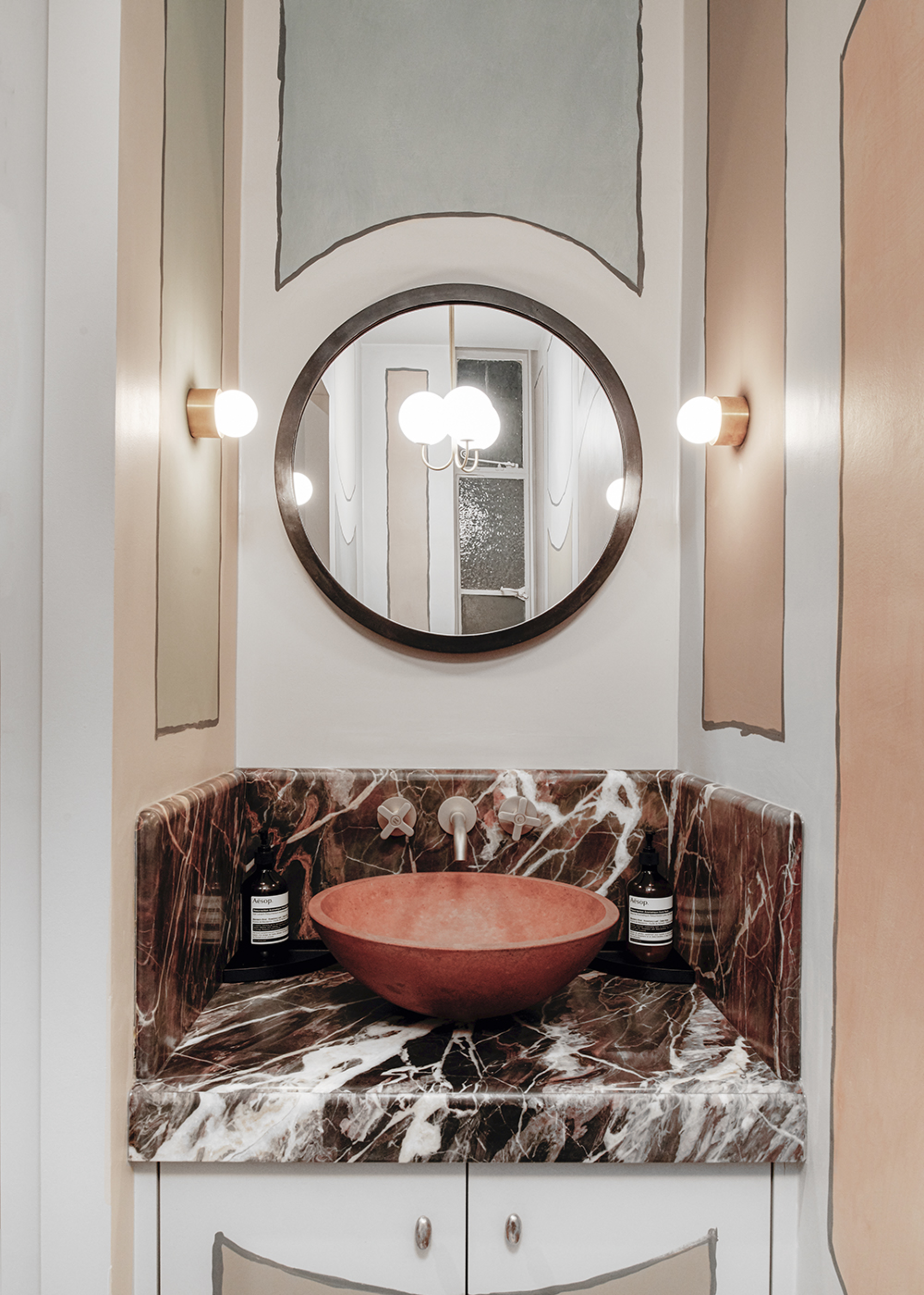 A powder room with brown veined marble vanity with a floating terracotta-hued sink, a round mirror above with wall lights either side and sketched lines adding a childlike artistic vibe