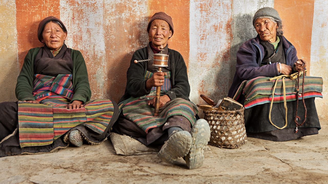 Three Tibetan people wearing traditional clothing sit against a wall