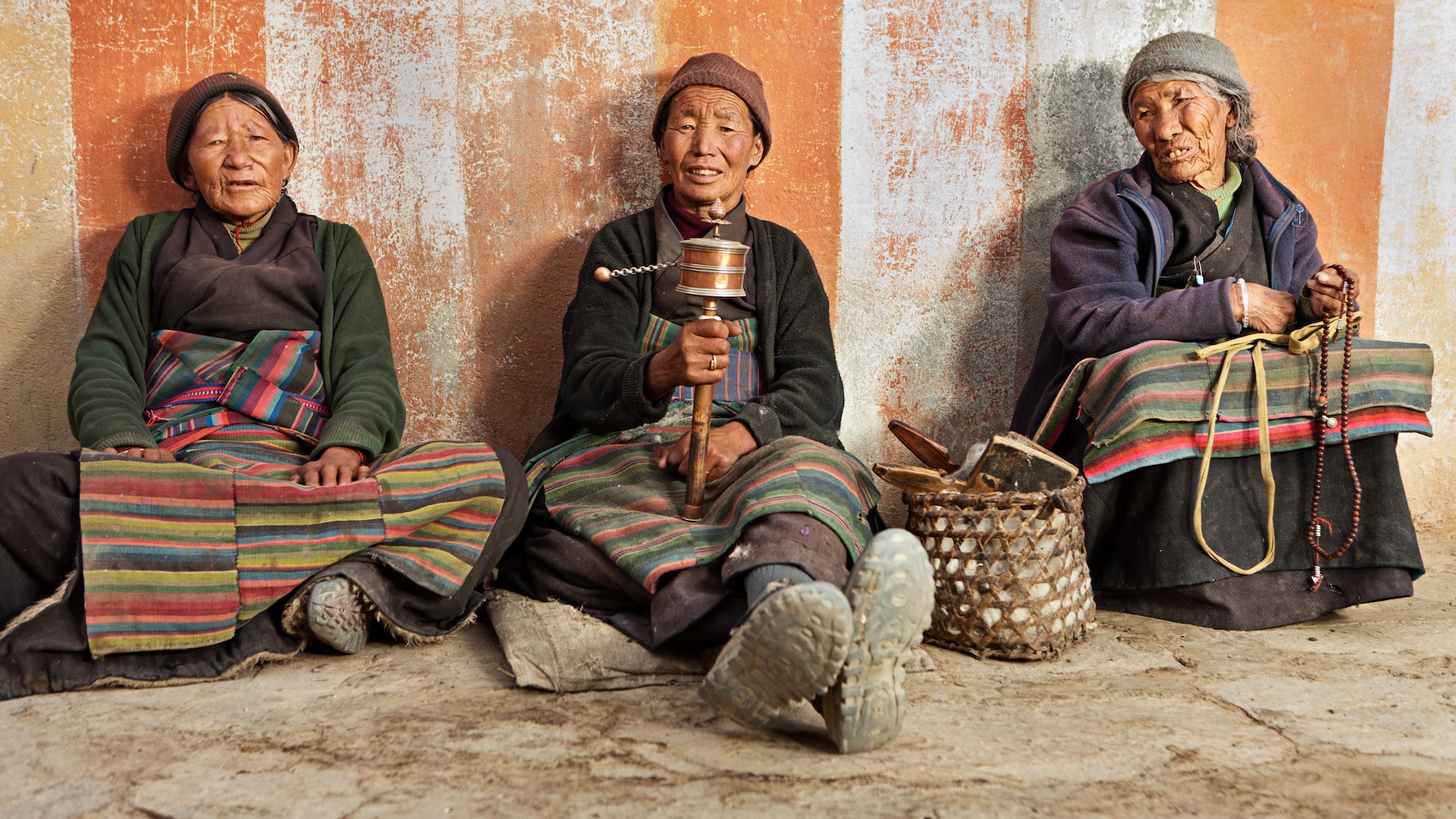Three Tibetan people wearing traditional clothing sit against a wall
