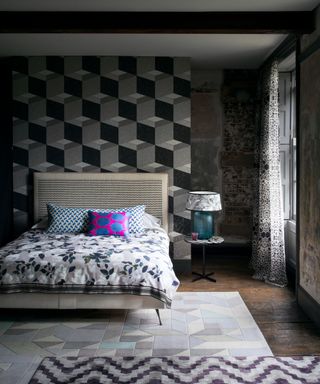 Geometric bedroom wallpaper in a dark, moody bedroom scheme with mismatched patterns including two different rug designs, floral bedlinen and spotty pillows.