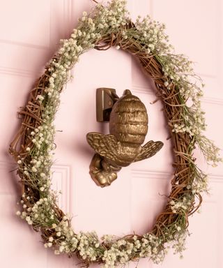 The pastel pink front door features an egg-shaped brown woven wreath with white flowers and a brushed brass bee door knocker