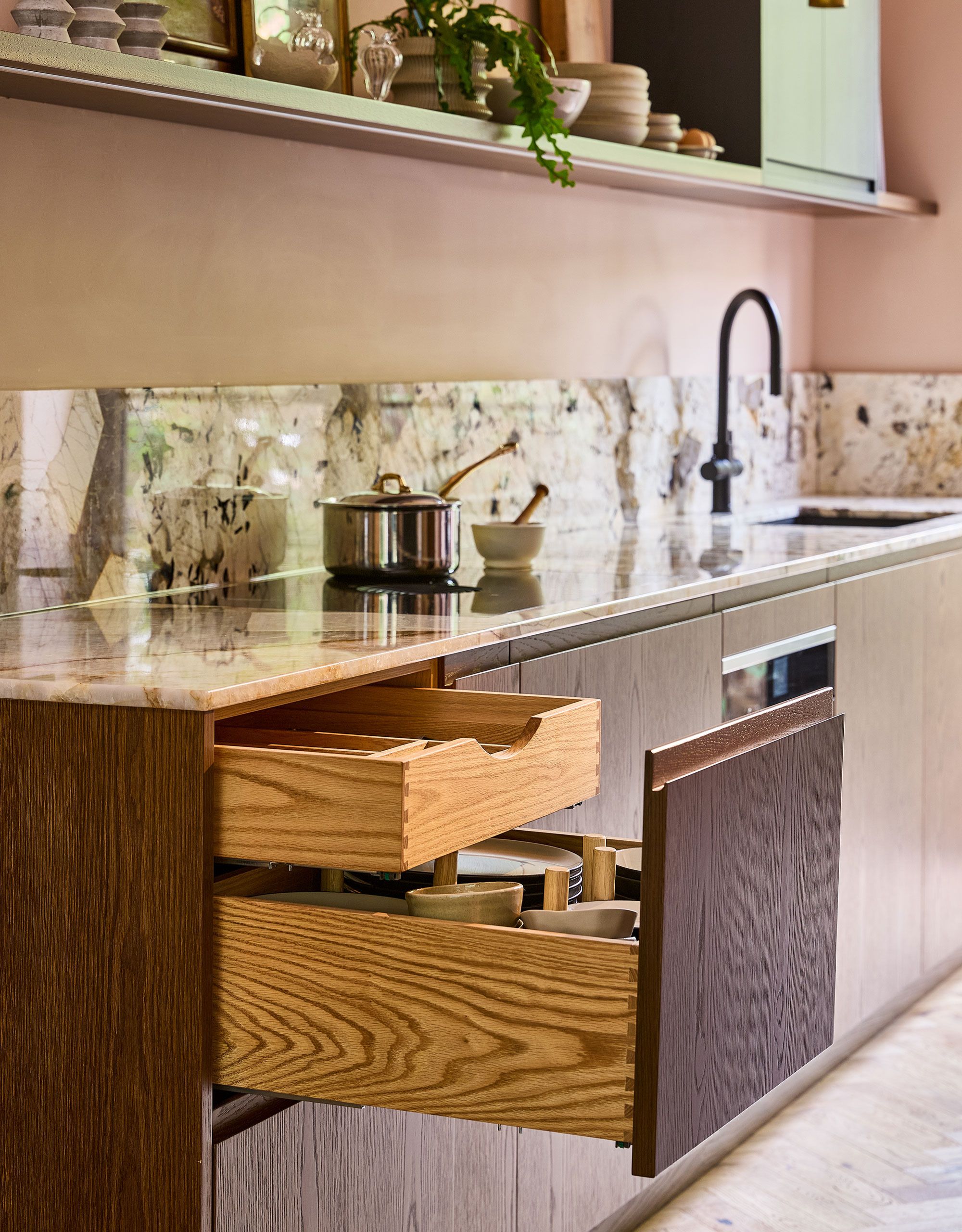 A modern kitchen with wood cabinetry and marble countertops and backsplash with pale pink ones, and one large kitchen drawer is open to expose two internal storage compartments