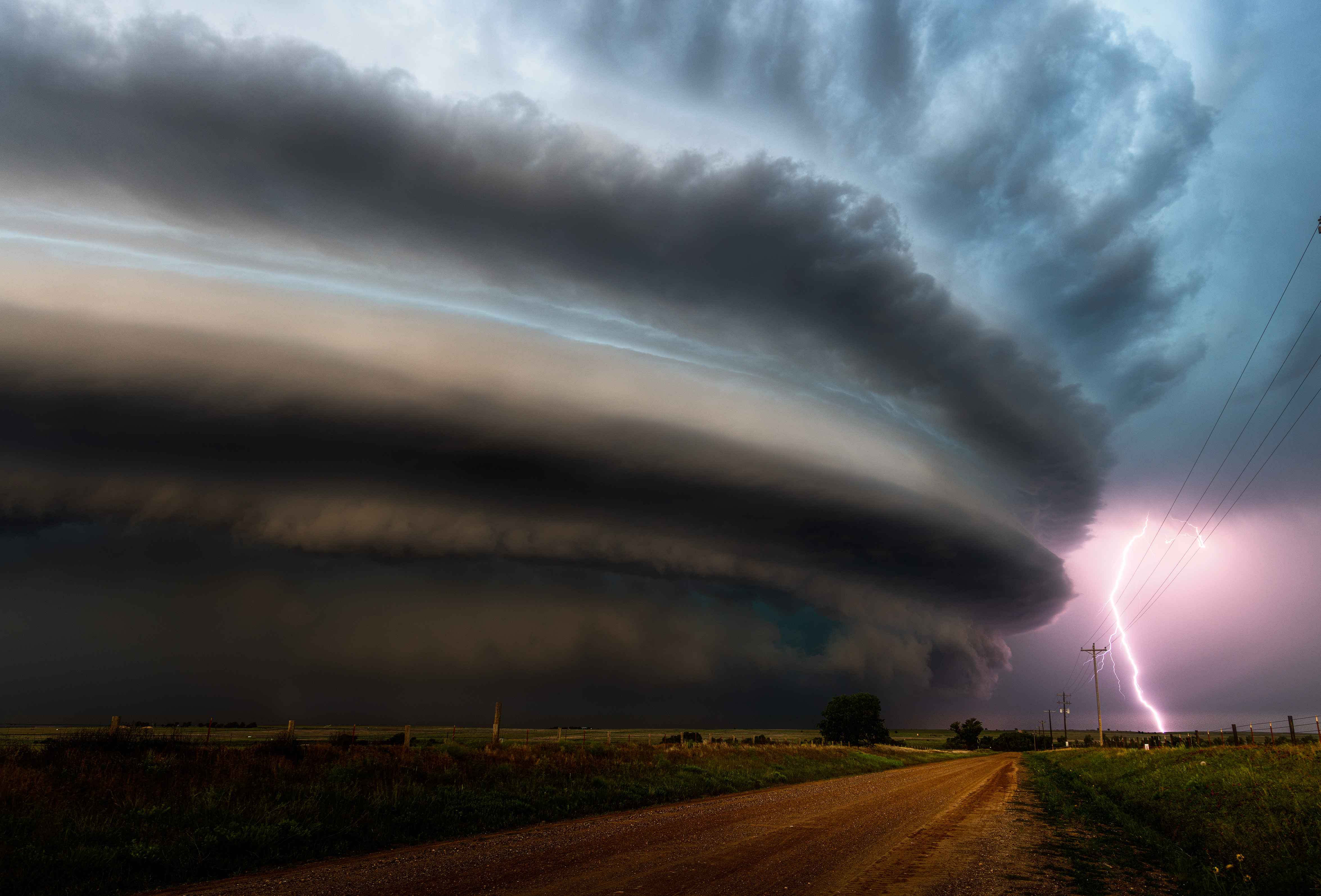 A powerful supercell thunderstorm with a massive, shelf-like cloud looms over a rural dirt road and field as a bolt of lightning strikes near a power pole.