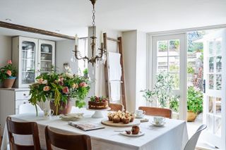 kitchen table with white cloth and wooden chairs and french window open candle chandelier and bouquet of flowers and cake on table