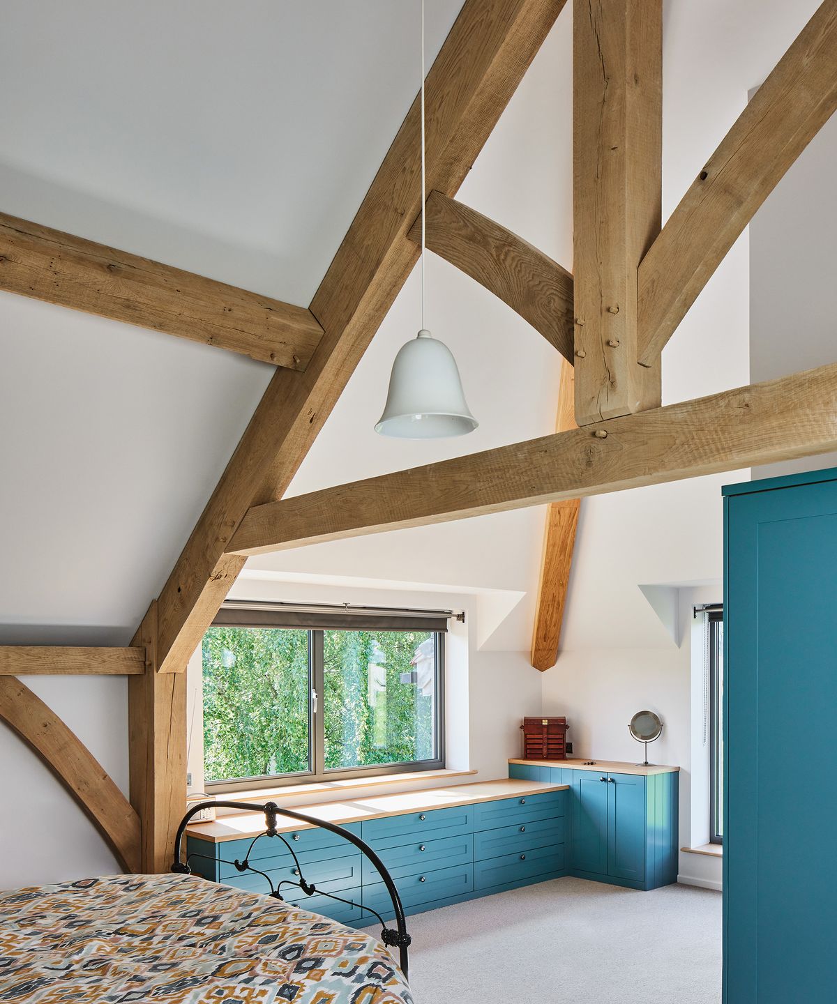 A bedroom with bespoke cabinetry in a bright teal colour. There are impressive exposed beams which form the frame. ofthe roof of this self build home