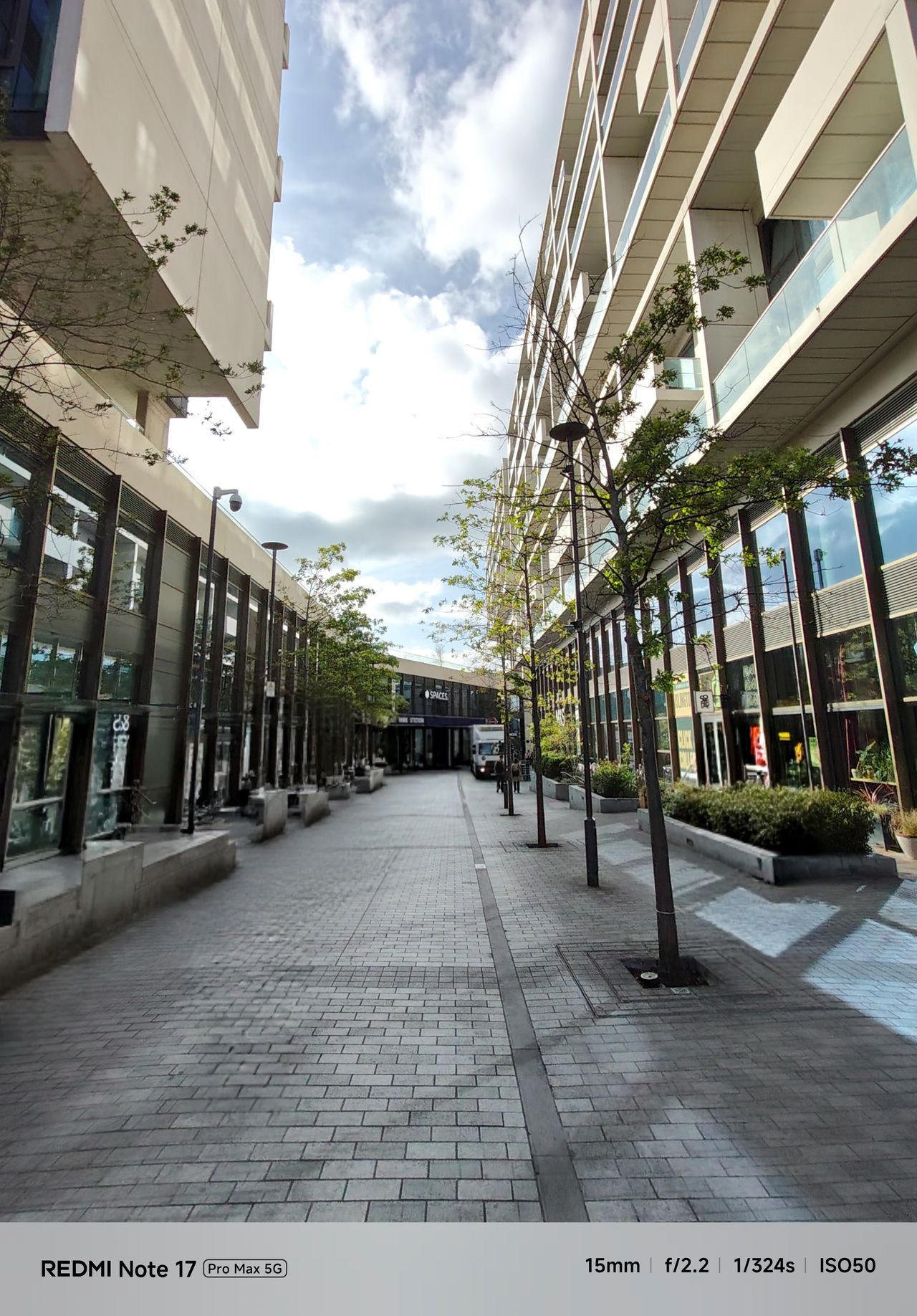 Wide-angle view down a pedestrian walkway with buildings, trees and a bright cloudy sky.