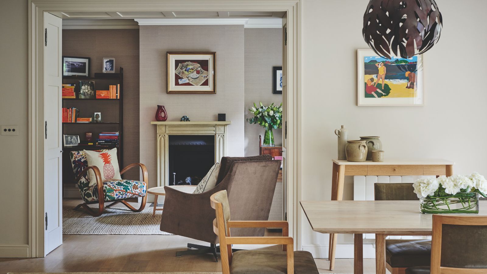 A neutral dining area with wooden chairs and table, and a doorway through to an eclectic neutral living room with upholstered chairs, a black fireplace with cream mantelpiece, artwork, flowers, and a black shelving unit filled with books.